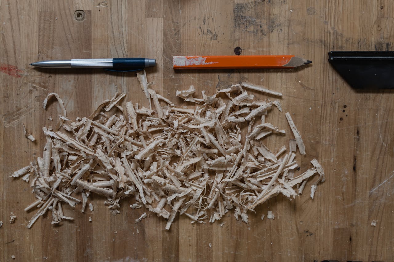 gallery-1 Close-up of wood shavings with a pen and a pencil on a wooden workbench, depicting craftsmanship.